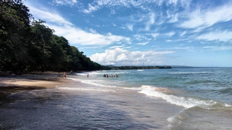 A beautiful beach in Costa Rica with clear blue water, a cloudy blue sky, and greenery.