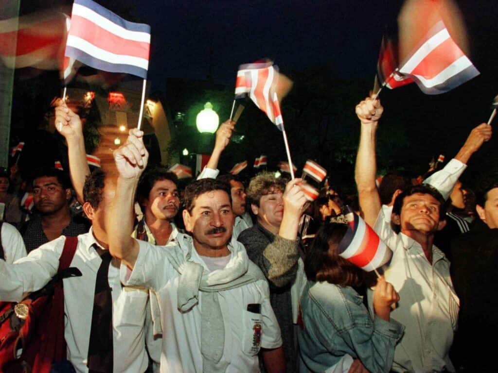 Several men walking in a parade at night waving small, paper Costa Rican flags.