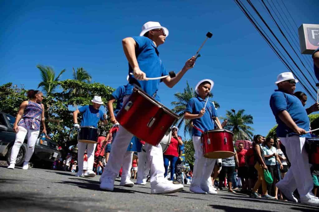 Several men in blue polos and white pants walking down a street playing red drums.