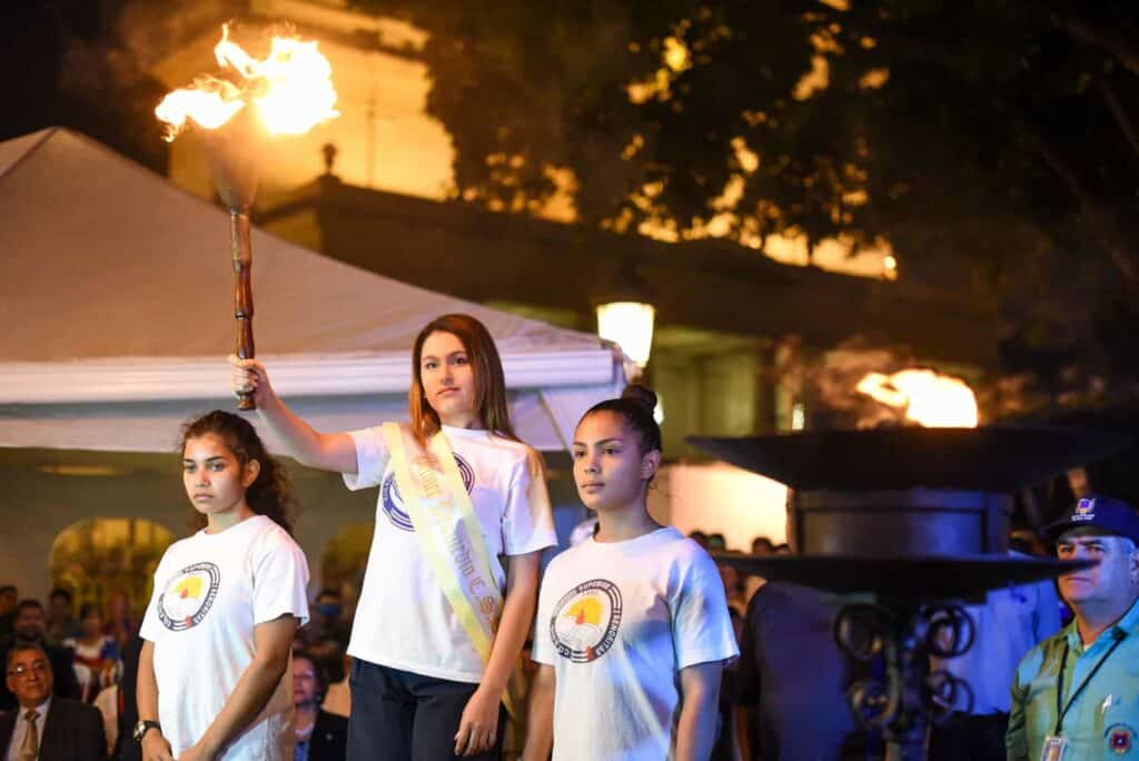 Three children in white shirts standing next to each other. One is holding a fiery torch in the air.
