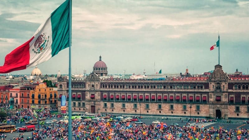 A large busy square in Mexico with several people milling about with the Mexican flag flying above it.