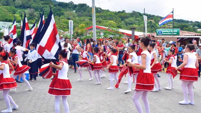 A parade in Costa Rica with several women and girls dressed in red skirts, white tights, and holding the Costa Rica flag.