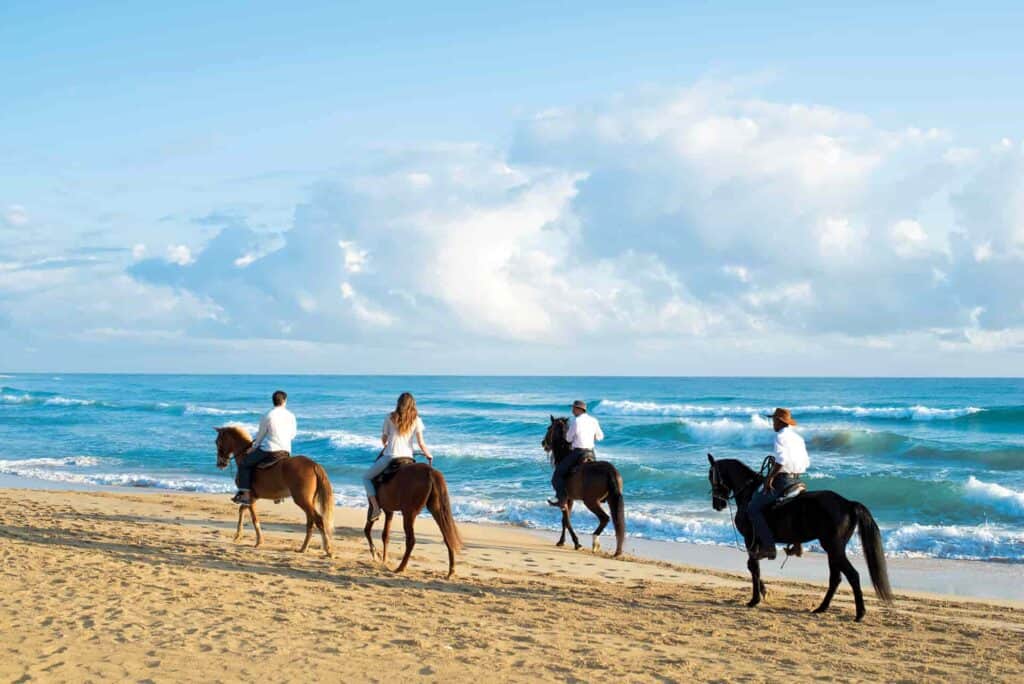 Four people in white shirts riding brown horses on a gold sand beach in the Dominican Republic