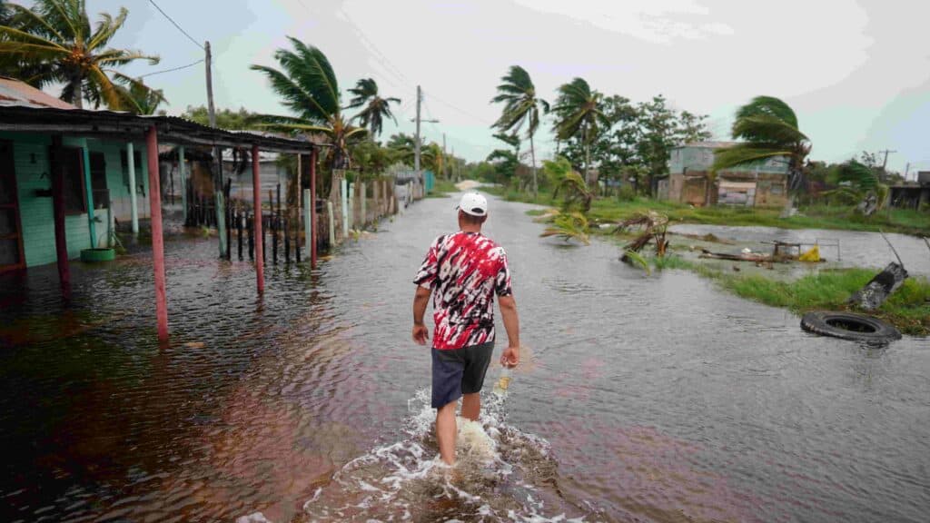 A man in a red, black, and white shirt and a white hat walking through an area in the Dominican Republic that was struck by a hurricane.