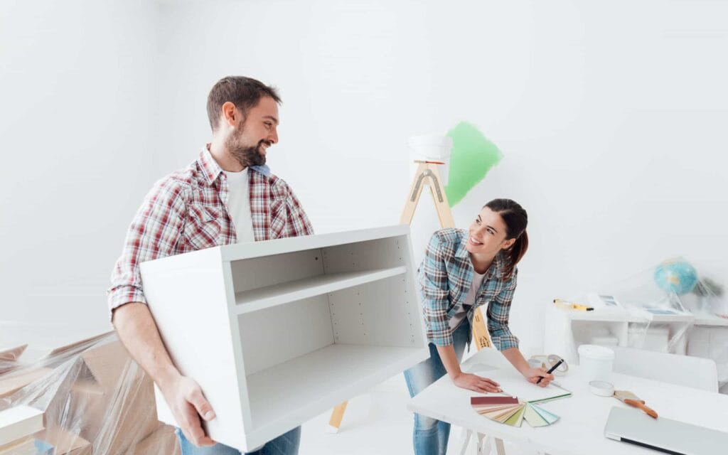 A couple in a white room with furniture and paint samples smiling at each other and working on home projects together.