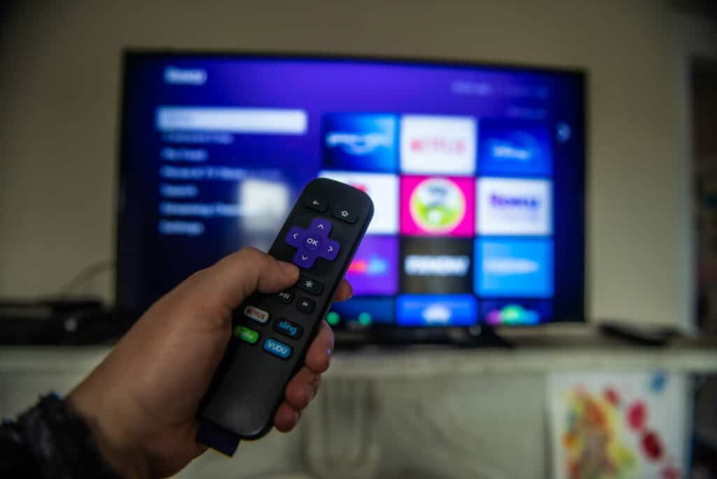 A Roku TV remote in front of a TV screen with someone pressing one of the buttons on the remote.