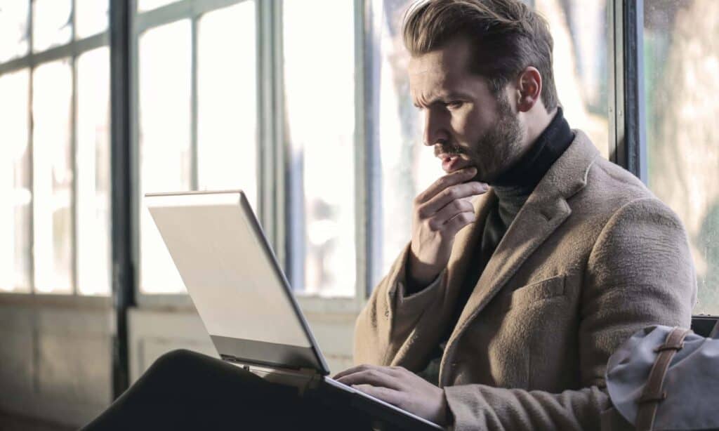 A man looking at a laptop computer screen stroking his beard with a contemplative look on his face.
