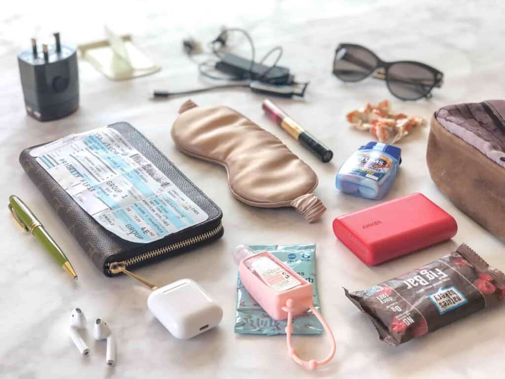 Several items from a carry-on bag laid on a bed, including a wallet, boarding pass, hand sanitizer, snack, tums, portable charger, sleep mask, sunglasses, AirPods, and more.