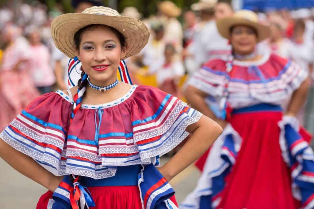 A woman standing in a traditional red, white, and blue dress and a straw hat during a parade during Costa Rican Independence Day.