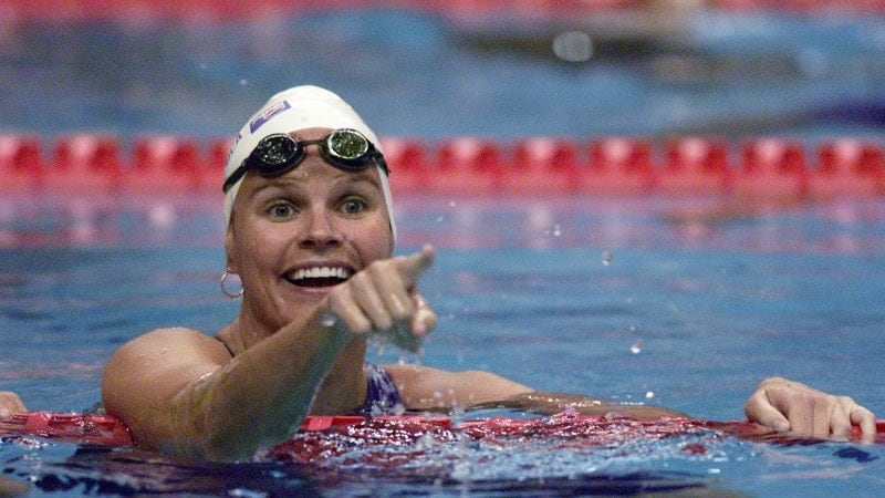 A woman pointing with her pointer finger in an Olympic swimming pool, wearing a white cap and black googles.