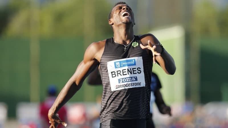 A man running across the finish line of a race in a black tank top with a look of distress on his face. He is wearing a white running bib that reads "Brenes"