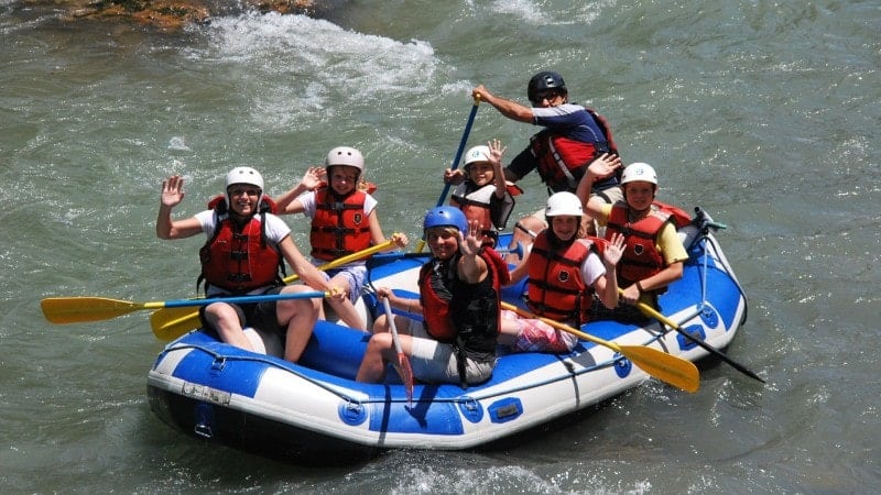 A group of seven adults and kids waving and wearing red vests on a white-water rafting tour.