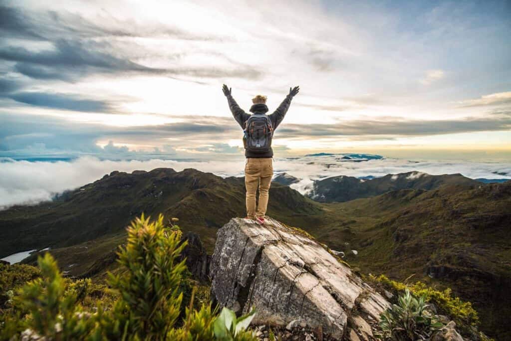 Person standing on the top of a mountain with their arms up in the air looking out at the view of the green mountains surrounding them.