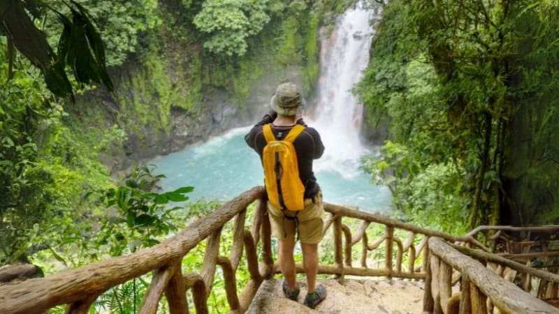a man taking a photo of a waterfall surrounded by green trees while hiking in Costa Rica