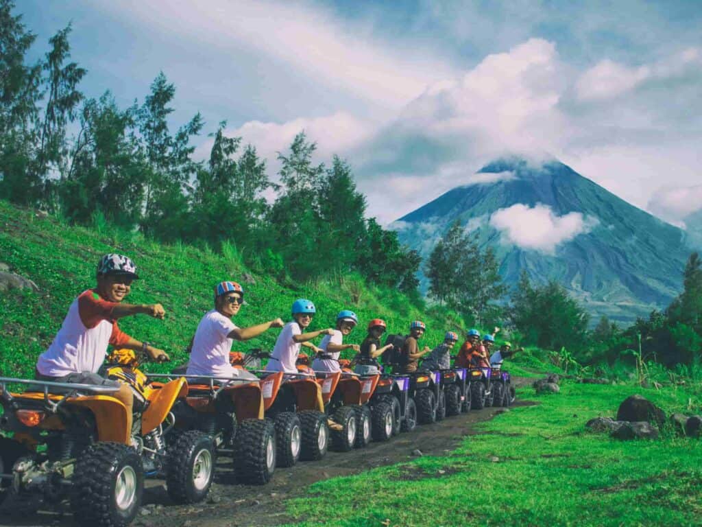 A line of men riding on ATVs while holding out a hand in a fist. The background is very green with lots of trees and a volcano.