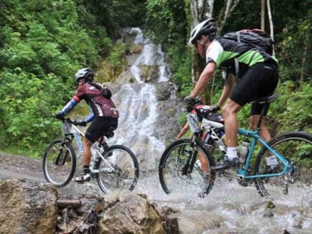 Three people riding up a mountain on mountain bikes in Costa Rica. There is a small waterfall in the the background.