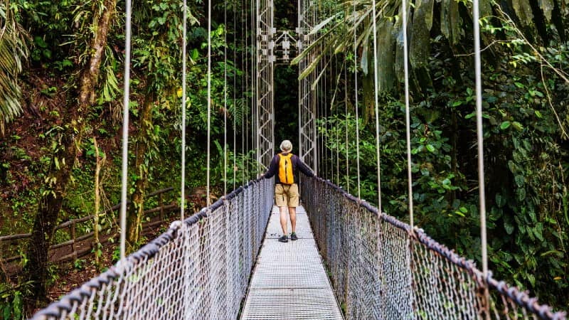 A man crossing a suspension bridge amidst lush jungle foliage.