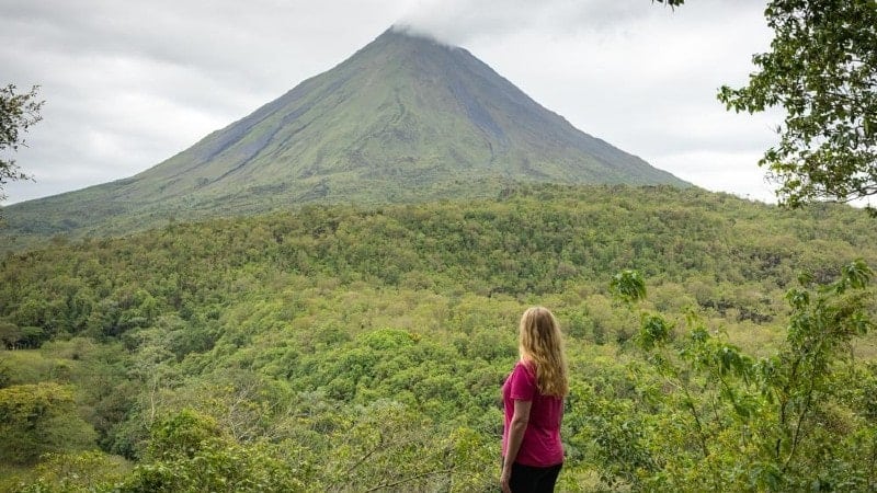 A woman in a pink shirt stands on a path, gazing at a mountain in the distance.