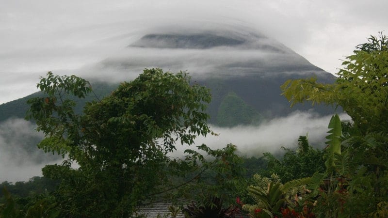 A misty volcano surrounded by lush vegetation and trees, concealed by a blanket of clouds.