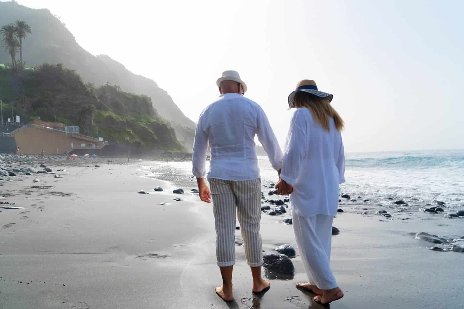 A retired couple holding hands and walking along a beach, with their backs to the camera. They are both wearing light, airy clothing and wide-brimmed hats. The beach is scattered with dark stones, and gentle waves lap at the shore. In the background, a green mountain slopes down to the coastline, and a building is nestled at the base of the slope, partially obscured by the foliage.