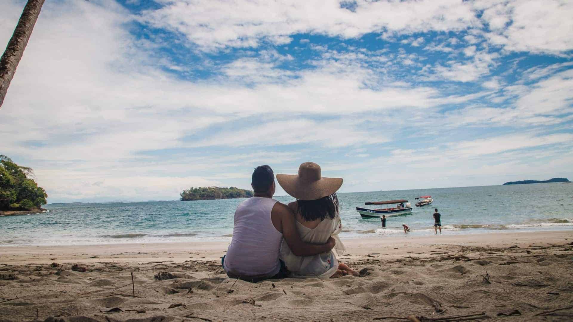 A couple sitting on the beach, gazing at the ocean waves with boats in the water.
