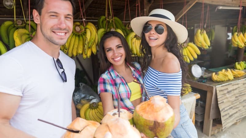 Three people smiling at the camera while holding coconuts. There are bananas in the background.