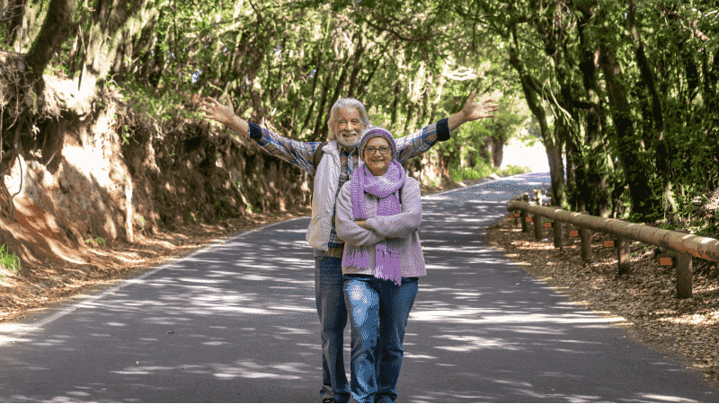 A joyful elderly couple stands in the middle of a tree-lined road, with the man's arms spread wide.