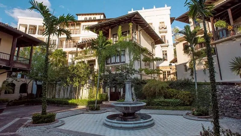 An elegant courtyard featuring a classic fountain at the center, surrounded by luxurious multi-story buildings with balconies adorned with lush hanging greenery. 