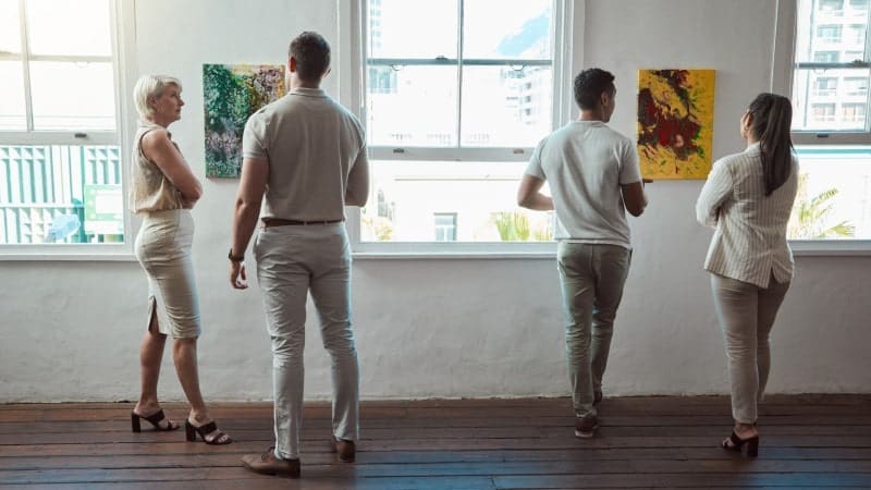 Four individuals are viewing artwork in a gallery with their backs to the camera. They are standing in front of a bright window. Two colorful paintings are hung on the wall.