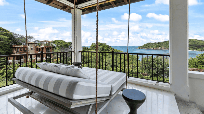 A luxurious hanging daybed on the balcony of a coastal home, offering a picturesque view of a lush, green coastline and the calm blue sea.