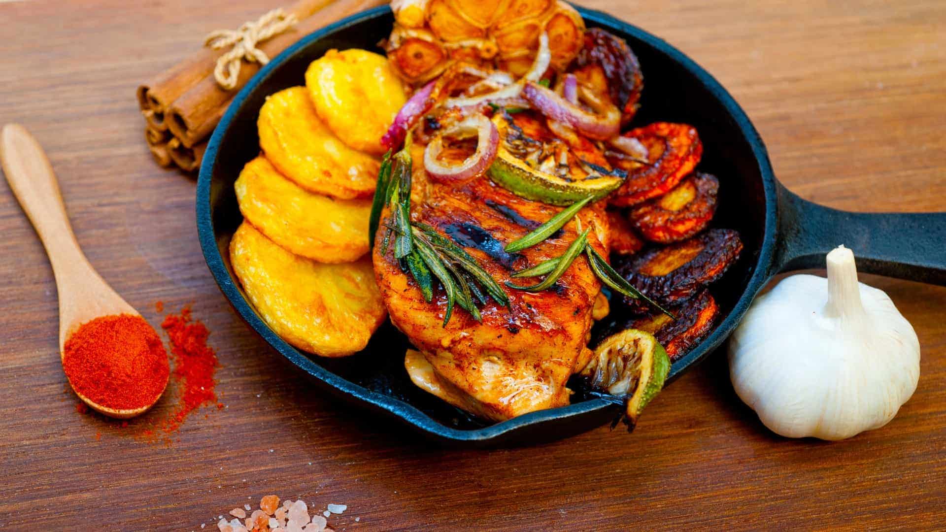 A vibrant cast-iron skillet filled with a juicy, grilled chicken breast, charred lime and red onion slices, and rosemary sprigs, accompanied by a side of golden fried plantains. The skillet is presented on a wooden table alongside a head of garlic, a spoonful of red ground spices, a pile of salt crystals, and cinnamon sticks.