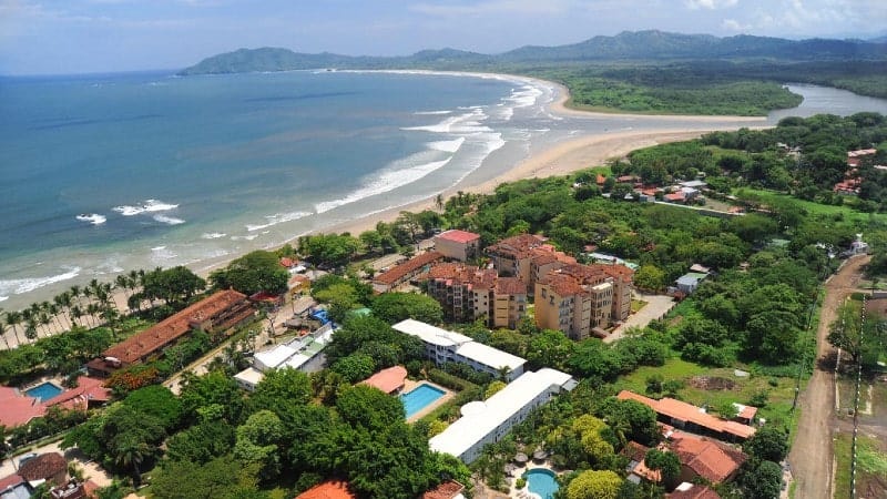 An aerial view of Tamarindo showcasing a coastal town with red-roofed buildings interspersed with lush greenery, multiple swimming pools, and nestled against a backdrop of a curving sandy beach.