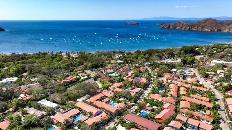 An aerial view of a beach town in Costa Rica, showcasing a vibrant community with rows of houses topped with red-tiled roofs, interspersed with lush greenery. 