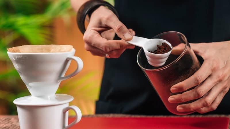 A person is using a spoon to transfer ground coffee from a brown glass container to a white ceramic pour-over coffee filter set on top of a cup.
