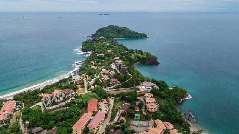 An aerial view of Playa Flamingo, Costa Rica, displays a lush peninsula with a mix of natural foliage and developed properties. The area features a combination of residential buildings with terracotta roofs, lush trees, and a curving shoreline edged by the clear turquoise waters of the Pacific Ocean. 