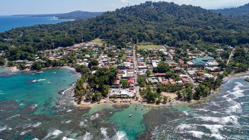 An aerial view of a vibrant beach town situated along the lush coast of Costa Rica. The town is a tapestry of colorful buildings, with a central street leading straight through the heart of the community toward the forested hills in the background.