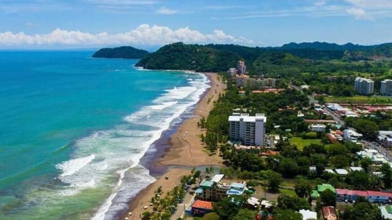 An aerial view of Jacó Beach in Costa Rica, featuring a long stretch of sandy shoreline with waves from the Pacific Ocean gently breaking on the beach. The coastal town is densely packed with a mix of residential and commercial buildings.