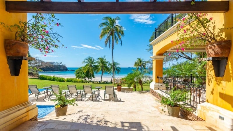 A view from a beachfront villa's shaded patio, looking out towards a picturesque tropical beach. 