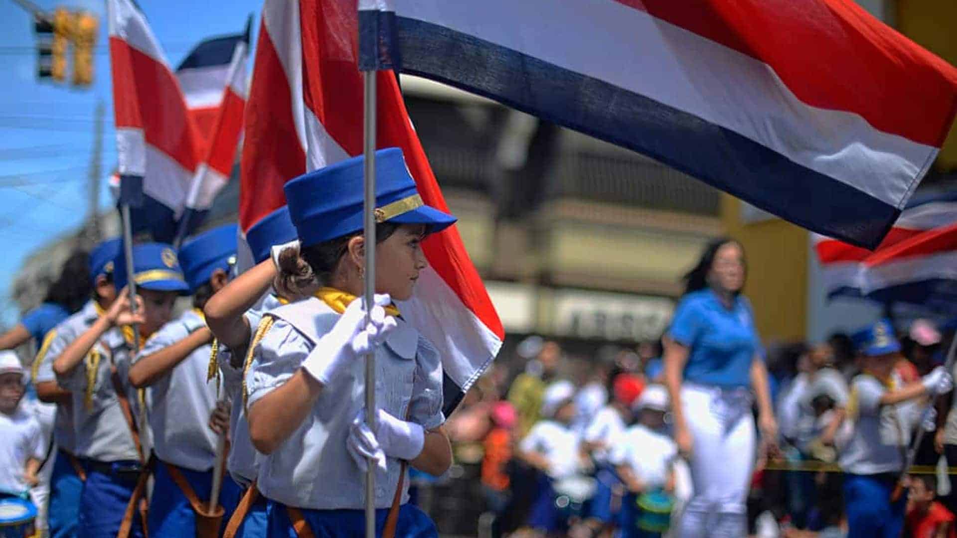 A group of children dressed in ceremonial uniforms, holding flags of Costa Rica, march in a parade. The prominent colors of the uniforms are blue and white, and the children also wear caps. The background is blurred with bystanders watching the event, emphasizing the children and flags in the foreground.
