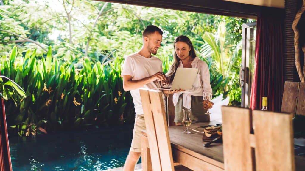 A couple is standing by a wooden counter in an open-air, tropical style kitchen with lush greenery visible in the background. The man, dressed casually in a white t-shirt and beige shorts, is holding a laptop and pointing at the screen, showing something to the woman next to him.