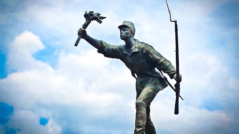A bronze statue of Juan Santamaría, a national hero of Costa Rica, holding a torch in his raised right hand and a rifle slung over his left shoulder. He is depicted mid-stride against a blue sky with white clouds. The statue commemorates his act of bravery during the Battle of Rivas in 1856.