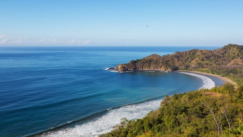 A panoramic view of a crescent-shaped bay with a sandy beach. The clear blue waters gently lap against the shore, bordered by lush greenery and rugged hills under a bright blue sky.