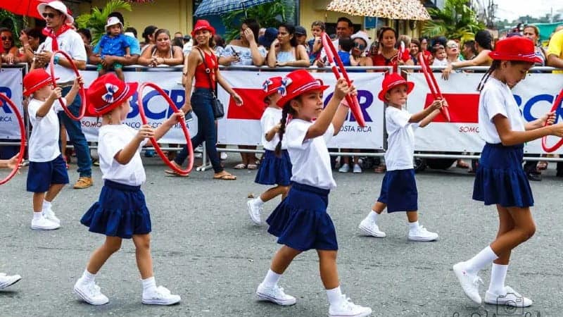 The image depicts a group of children dressed in uniforms, participating in a parade. They are wearing white shirts, navy blue skirts, and red hats, marching in step while holding small red hoops. The background is filled with spectators and banners, indicating a festive, public event. 