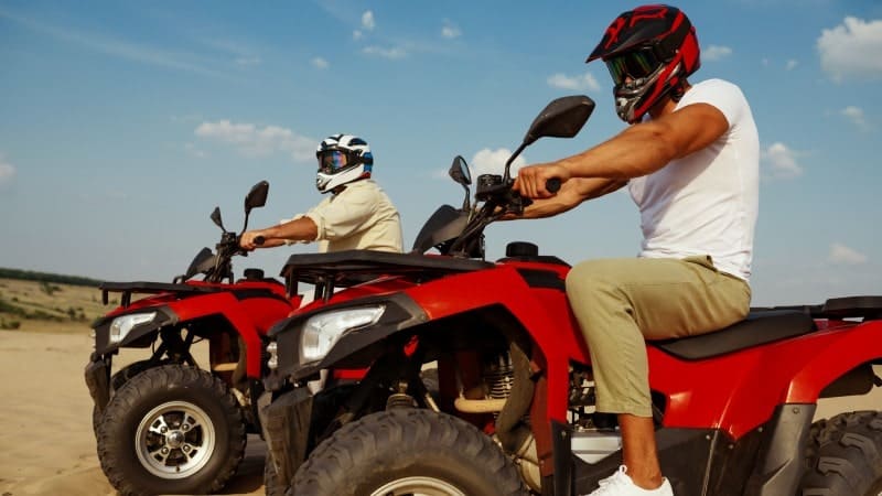 Two individuals riding red all-terrain vehicles (ATVs) across a sandy area under a clear sky, with both riders wearing protective helmets.