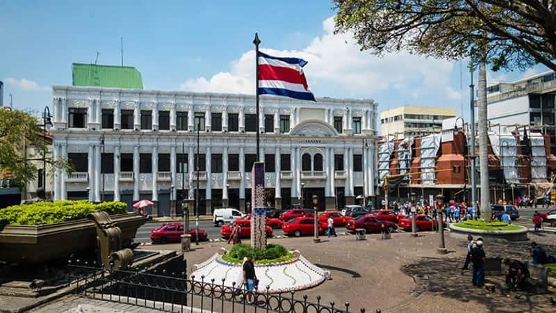 The image features an urban scene with a grand, white neoclassical building featuring an arched central entrance and multiple windows. A Costa Rican flag flies prominently in front of the building. In the foreground, there's a busy street with red cars and a few pedestrians.