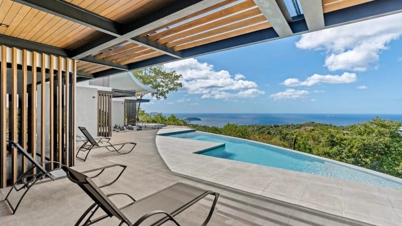 The image showcases a modern terrace with a luxurious infinity pool overlooking a stunning coastal view. The pool's edge seamlessly merges with the ocean horizon. The terrace is adorned with comfortable sun loungers, and the design features a combination of wooden slats and beams that create a contemporary, open-air structure. Lush greenery surrounds the area.