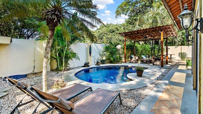 A private, serene backyard oasis featuring a round blue swimming pool surrounded by lush tropical greenery and a variety of plants, including a prominent palm tree. To the left, two sun loungers invite relaxation under the sun on a pebble-covered area, and to the right, a covered pergola with outdoor furniture suggests a space for dining or lounging in the shade. The yard is enclosed by a high white wall, providing privacy, and a classic lantern-style light fixture adds to the tranquil ambiance. 