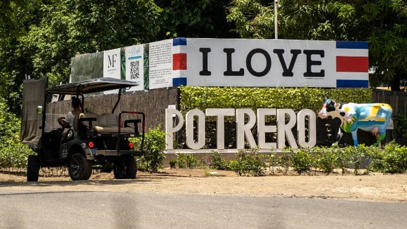 The image features a roadside sign that reads "I LOVE POTRERO" in large, bold letters. Adjacent to the sign, there's a colorful, cow-shaped statue painted with various designs, adding a playful touch to the scene. A golf cart with a person driving it is parked on the left side of the image.