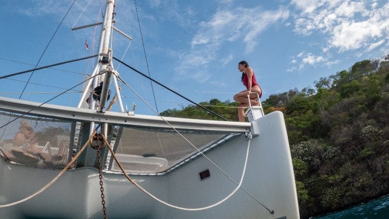 A woman stands on the bow of a sailboat, looking out towards the sea. She's poised near the vessel's prow, leaning slightly forward with her hands on the railings.