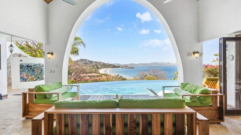 View from an elegant archway of a luxurious seaside villa, featuring a tranquil infinity pool that blends with the panoramic vista of a bay. Plush green cushions adorn wooden patio furniture, inviting relaxation with a scenic backdrop.